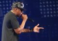 Jay-Z in a black cap, sunglasses and gold chain raps into a microphone and points in front of a blue-lit wall of stage lights