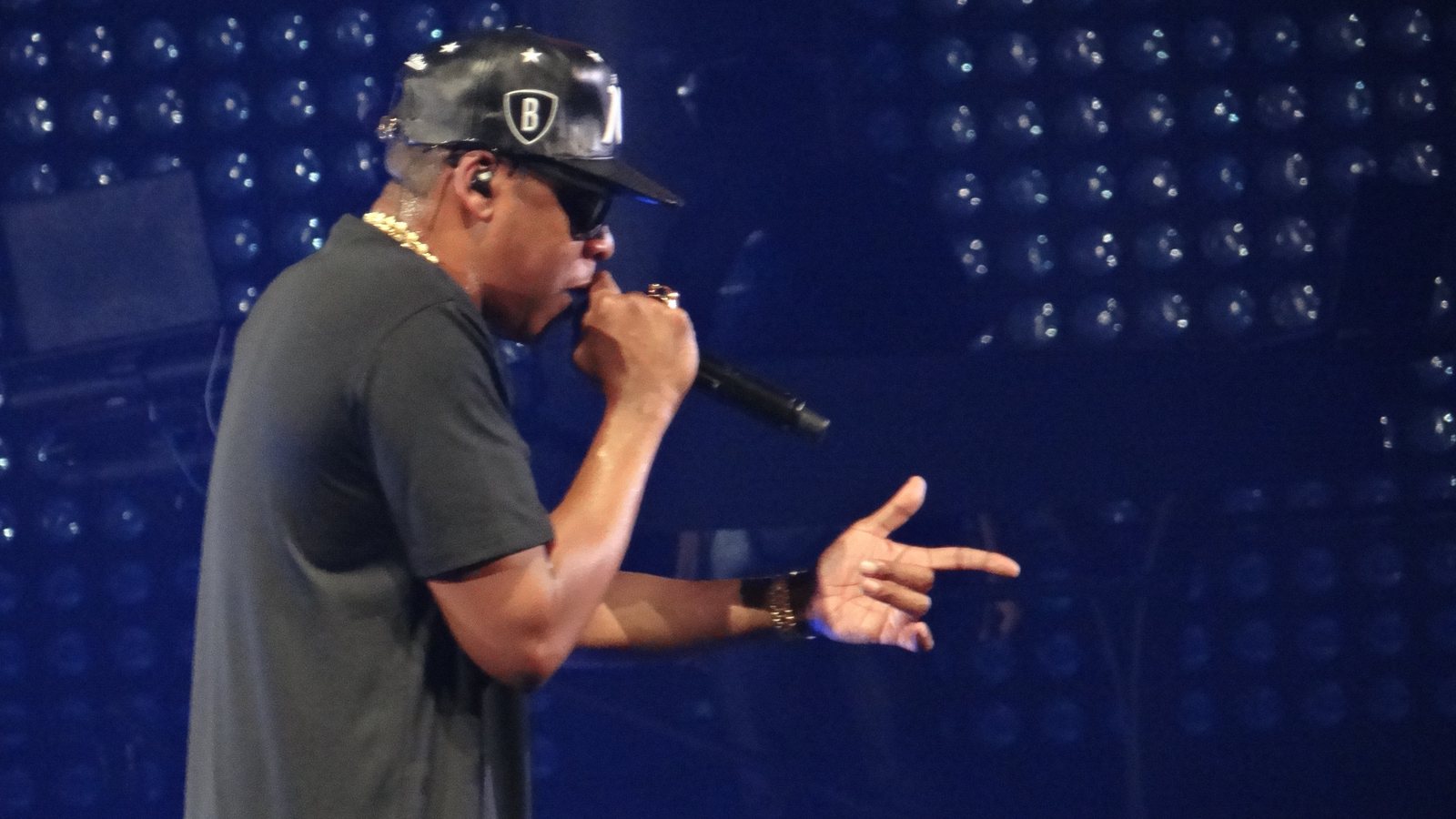 Jay-Z in a black cap, sunglasses and gold chain raps into a microphone and points in front of a blue-lit wall of stage lights