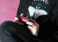 A person in a black butterfly T-shirt sits on a pink couch holding a pink PlayStation 5 DualSense controller with both hands