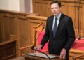 James Comey in a dark suit and blue tie speaking from a wooden lectern with a microphone, red chairs and wood paneling behind him