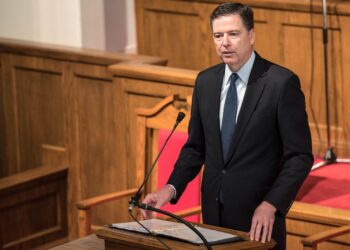 James Comey in a dark suit and blue tie speaking from a wooden lectern with a microphone, red chairs and wood paneling behind him