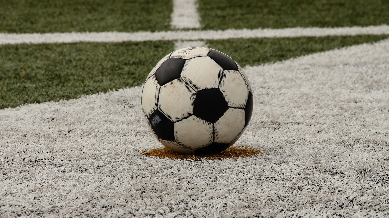 A worn black-and-white soccer ball resting on a painted spot on an artificial turf field