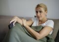 A woman wrapped in a green blanket sits on a gray couch holding a TV remote control