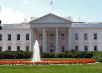 The north facade of the White House in Washington, with its columned portico, and a fountain on the North Lawn in front, on a clear day