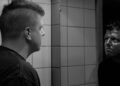 A man with a short mohawk haircut studies his reflection in a bathroom mirror, in black and white
