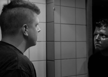 A man with a short mohawk haircut studies his reflection in a bathroom mirror, in black and white