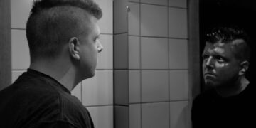 A man with a short mohawk haircut studies his reflection in a bathroom mirror, in black and white