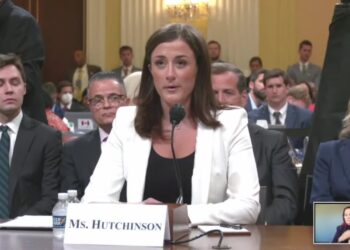 Cassidy Hutchinson in a white jacket seated at a witness table behind a microphone and a name card reading Ms. Hutchinson, with people seated behind her