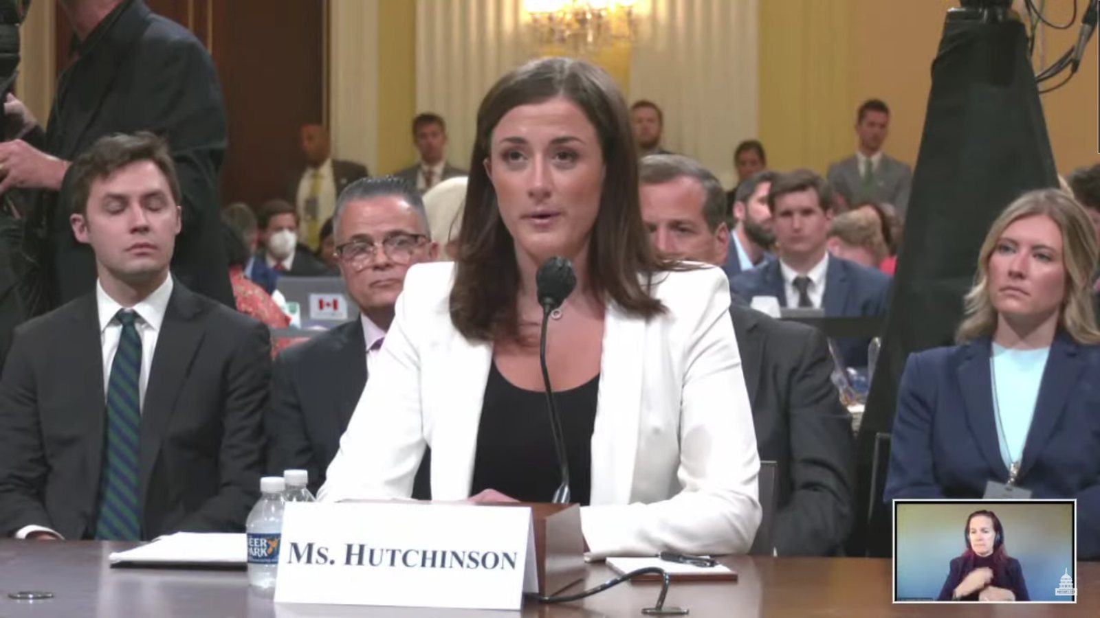 Cassidy Hutchinson in a white jacket seated at a witness table behind a microphone and a name card reading Ms. Hutchinson, with people seated behind her