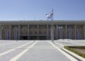 The front of the Knesset building in Jerusalem under a clear blue sky, with Israeli flags on a pole in the plaza and banners on the facade