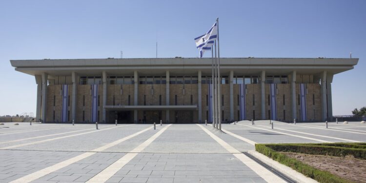 The front of the Knesset building in Jerusalem under a clear blue sky, with Israeli flags on a pole in the plaza and banners on the facade