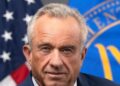 Health Secretary Robert F. Kennedy Jr. in his official portrait, in a dark pinstripe suit in front of the US flag and the HHS flag