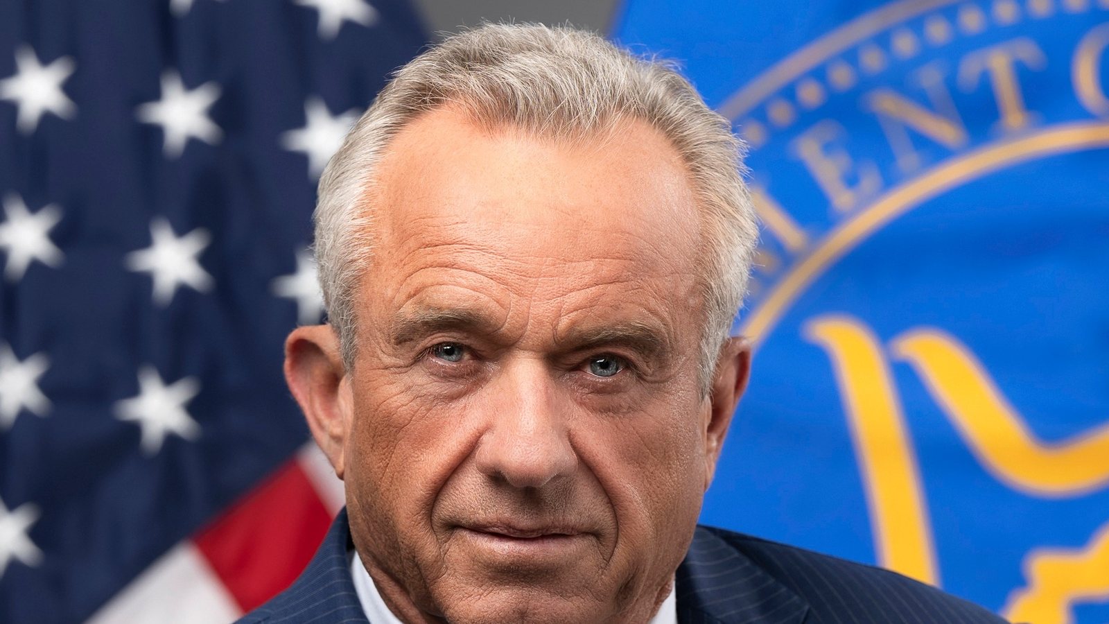 Health Secretary Robert F. Kennedy Jr. in his official portrait, in a dark pinstripe suit in front of the US flag and the HHS flag