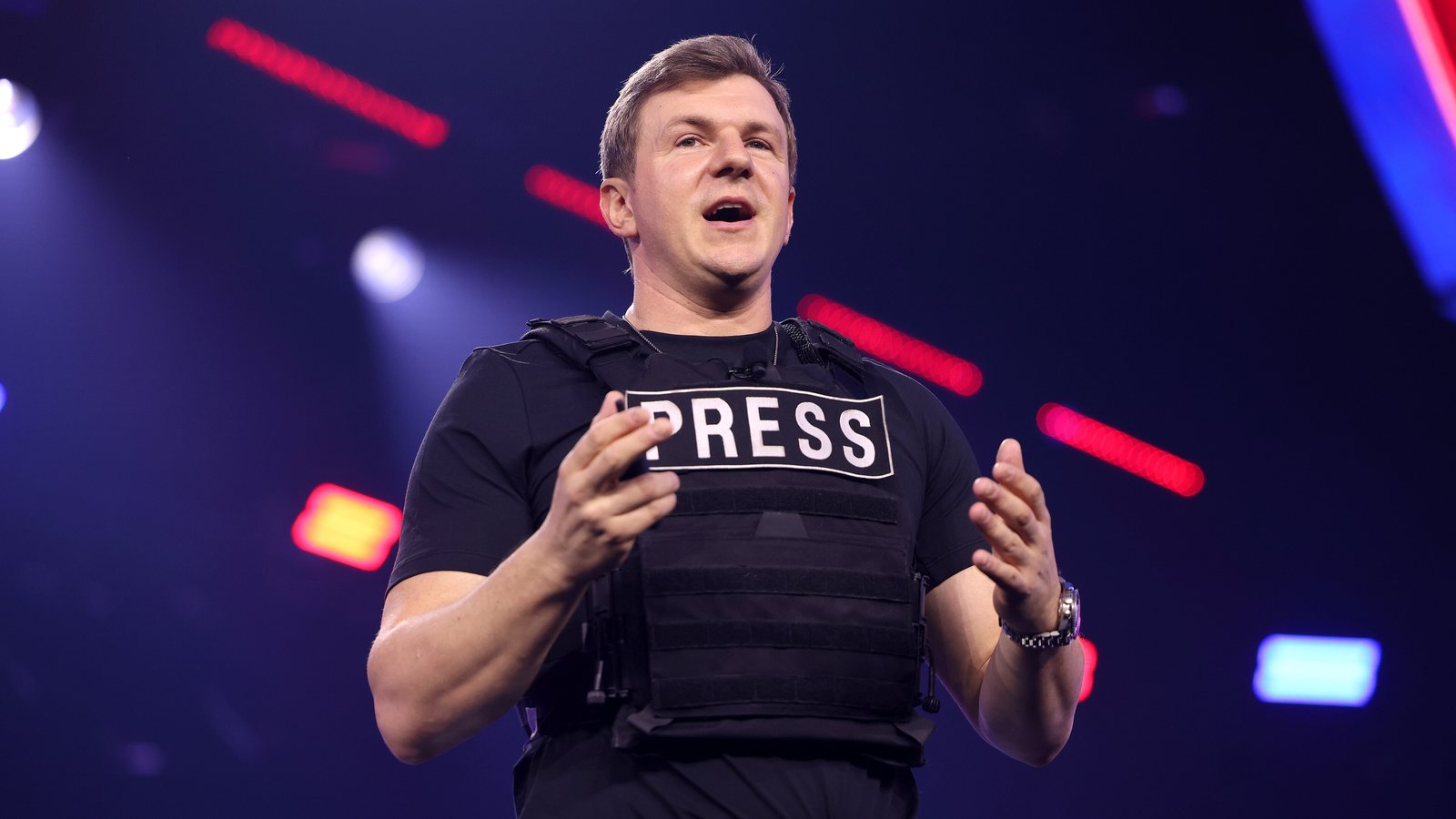 James O'Keefe in a black t-shirt and a vest marked PRESS speaking on a brightly lit stage