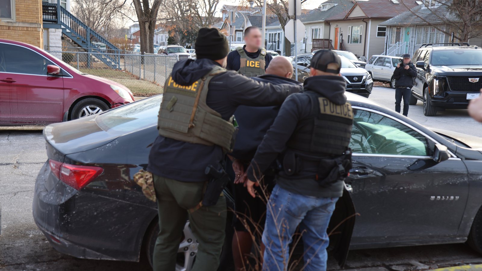 ICE officers in vests marked Police and ICE, seen from behind, escort a man to a car on a residential street in Chicago