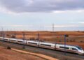 A white Renfe AVE Class 103 high-speed train runs through dry farmland under overhead power lines