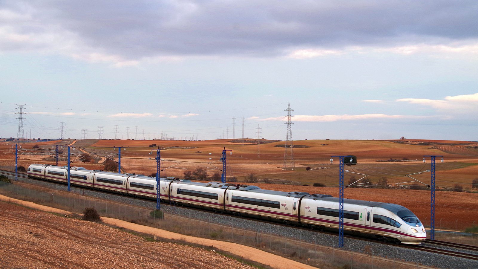 A white Renfe AVE Class 103 high-speed train runs through dry farmland under overhead power lines