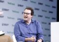 Anthropic chief executive Dario Amodei, in glasses and a blue shirt, speaks from a white armchair on the TechCrunch Disrupt stage