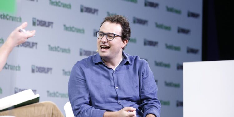 Anthropic chief executive Dario Amodei, in glasses and a blue shirt, speaks from a white armchair on the TechCrunch Disrupt stage