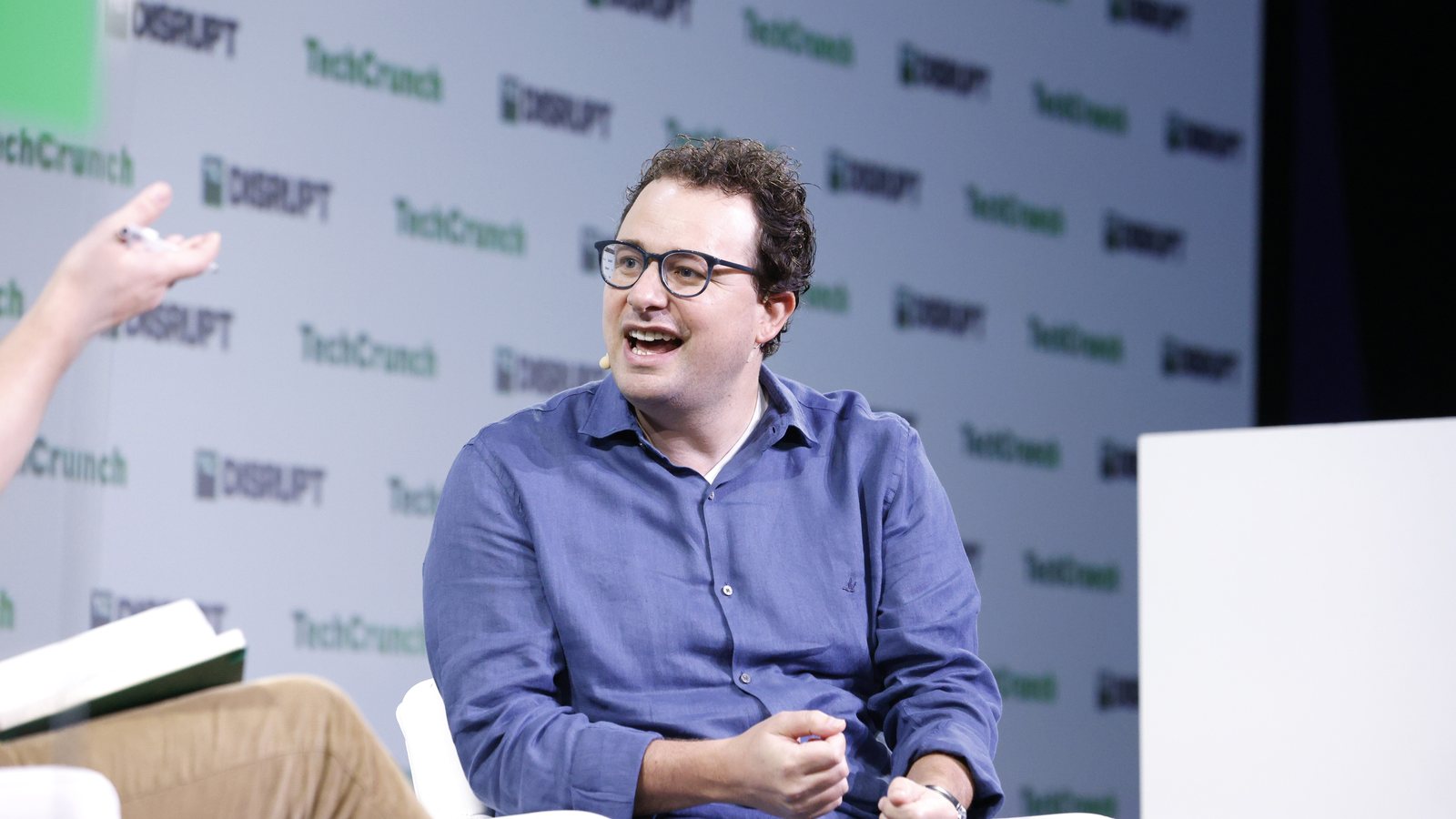Anthropic chief executive Dario Amodei, in glasses and a blue shirt, speaks from a white armchair on the TechCrunch Disrupt stage