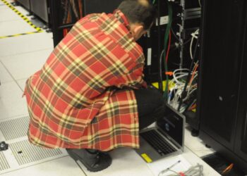 A technician in a plaid shirt crouches with a laptop beside an open server rack in a data center