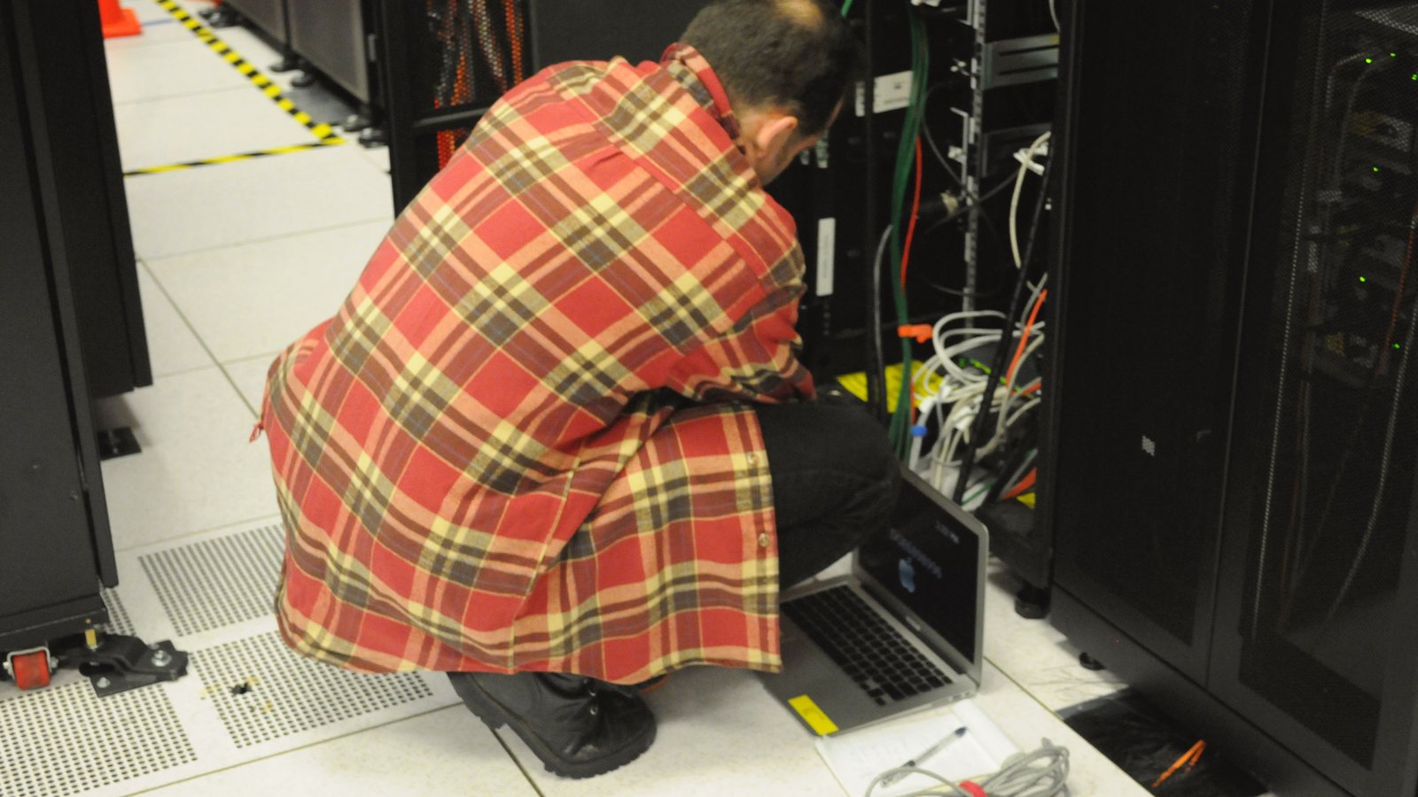A technician in a plaid shirt crouches with a laptop beside an open server rack in a data center