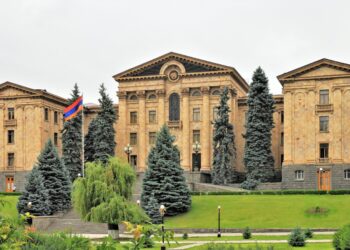 The National Assembly building in Yerevan, Armenia, a long stone facade with columns and a flag, behind fir trees and a lawn
