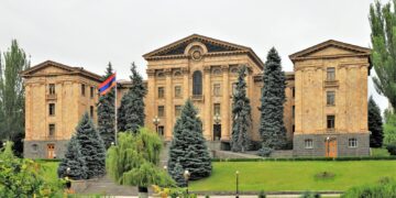 The National Assembly building in Yerevan, Armenia, a long stone facade with columns and a flag, behind fir trees and a lawn