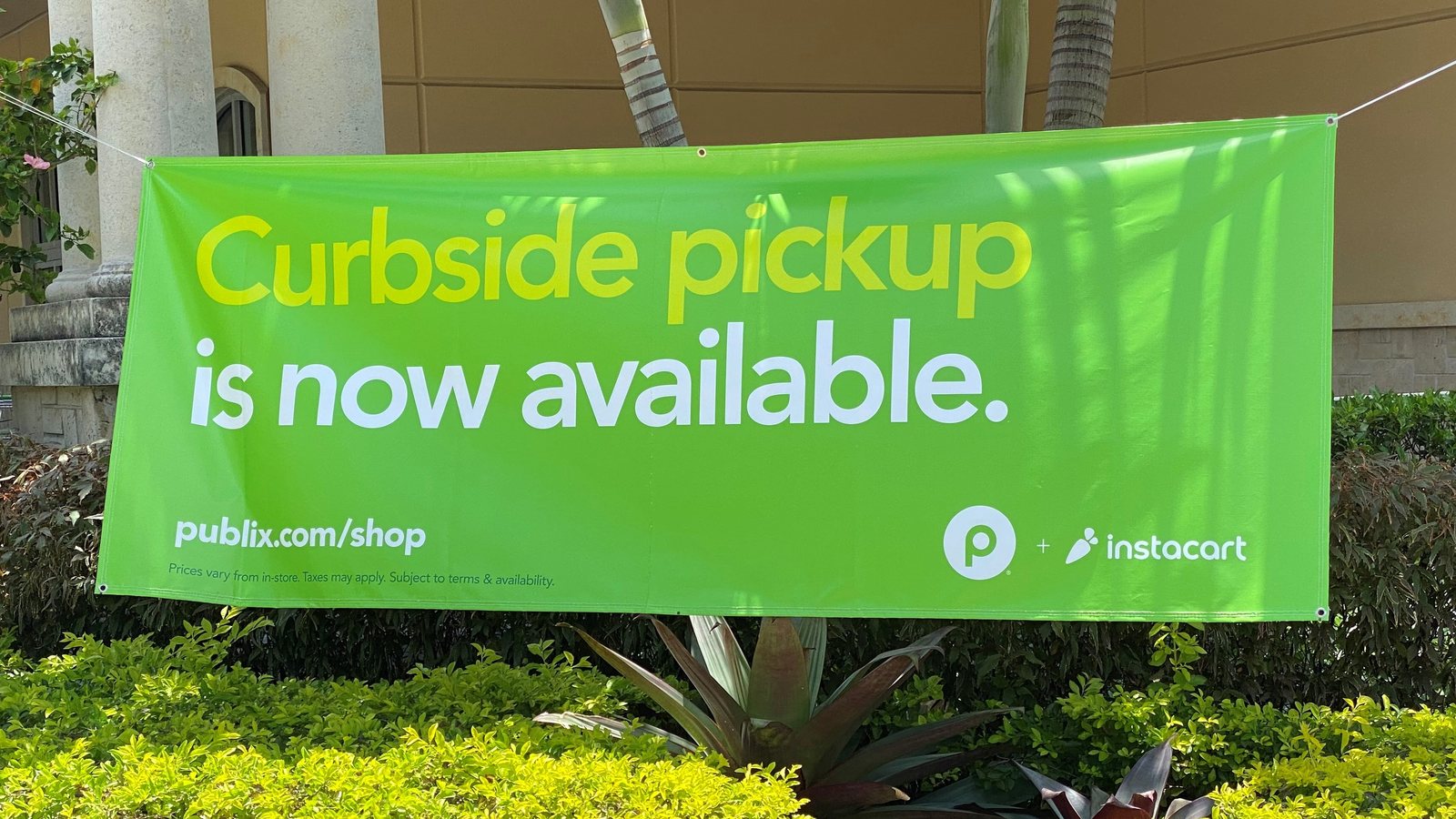 A green Publix and Instacart banner reading Curbside pickup is now available, hung above shrubs outside a store in West Miami