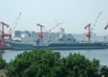 A gray aircraft carrier docked beside orange shipyard cranes, with apartment towers behind it