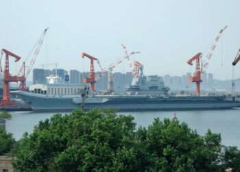 A gray aircraft carrier docked beside orange shipyard cranes, with apartment towers behind it