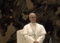 Pope Leo XIV in white papal vestments and pectoral cross, standing and smiling in front of a bronze sculpture in the Vatican's audience hall