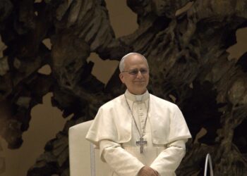 Pope Leo XIV in white papal vestments and pectoral cross, standing and smiling in front of a bronze sculpture in the Vatican's audience hall