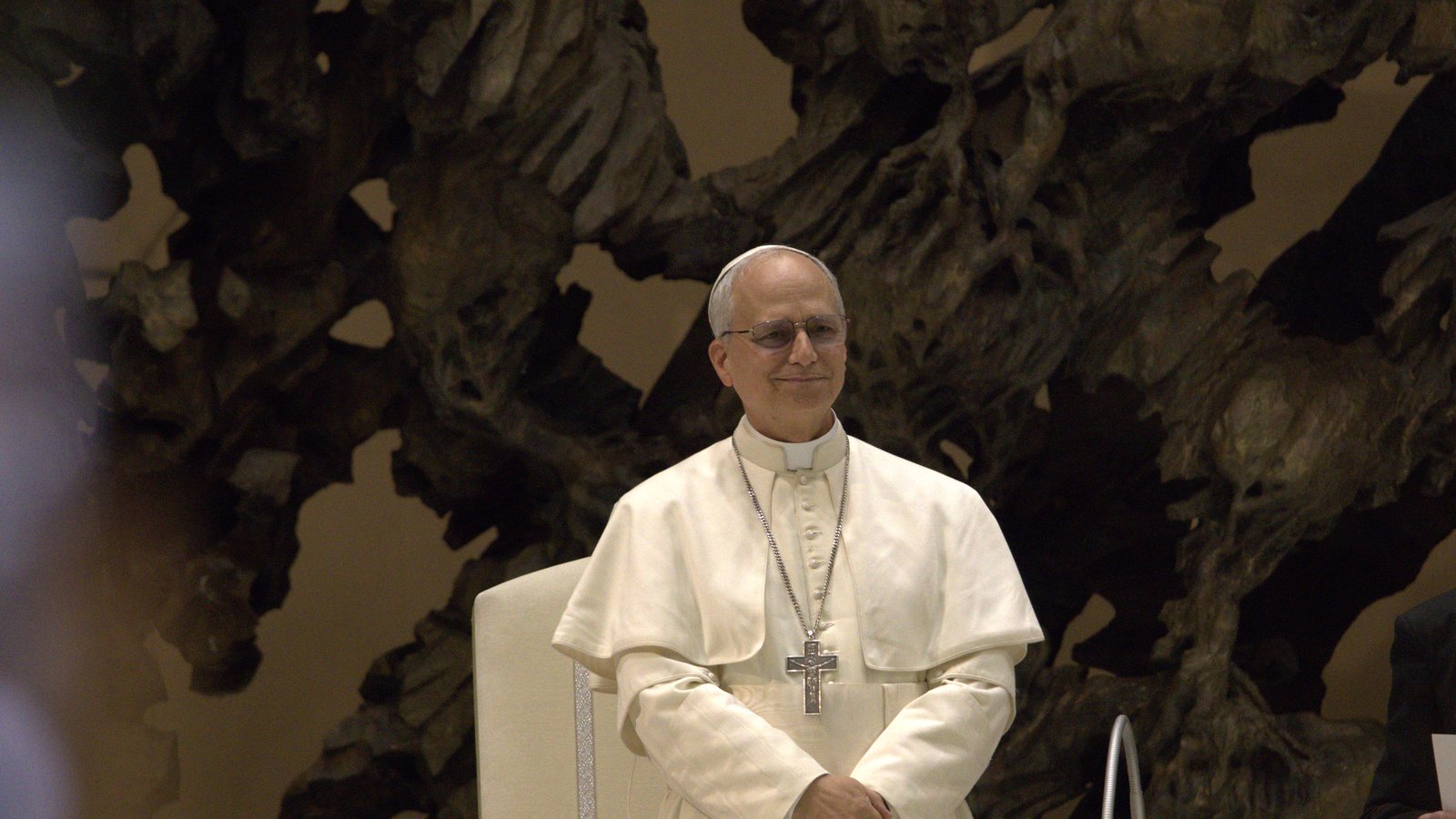 Pope Leo XIV in white papal vestments and pectoral cross, standing and smiling in front of a bronze sculpture in the Vatican's audience hall