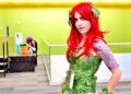 A cosplayer dressed as Poison Ivy, with long red hair and a green leaf costume, posing at a fan convention