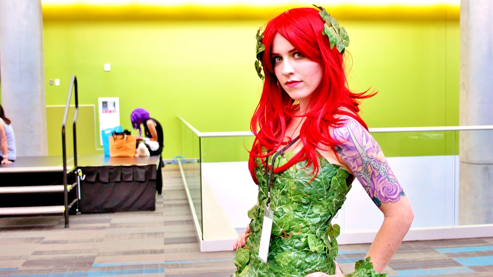 A cosplayer dressed as Poison Ivy, with long red hair and a green leaf costume, posing at a fan convention