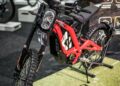 A red Sur-Ron electric dirt bike with foot pegs and no pedals on display at a trade show