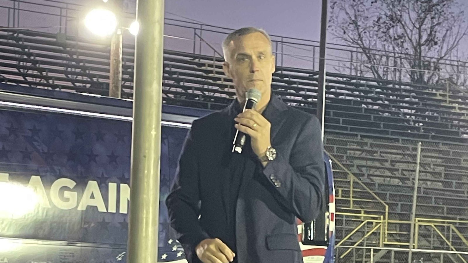 Corey Lewandowski holds a microphone while speaking in front of a Trump campaign bus at a speedway at dusk