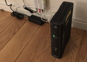 An AT&T BGW210 Wi-Fi gateway standing on a wood floor next to a wall outlet, with its WPS button on the front