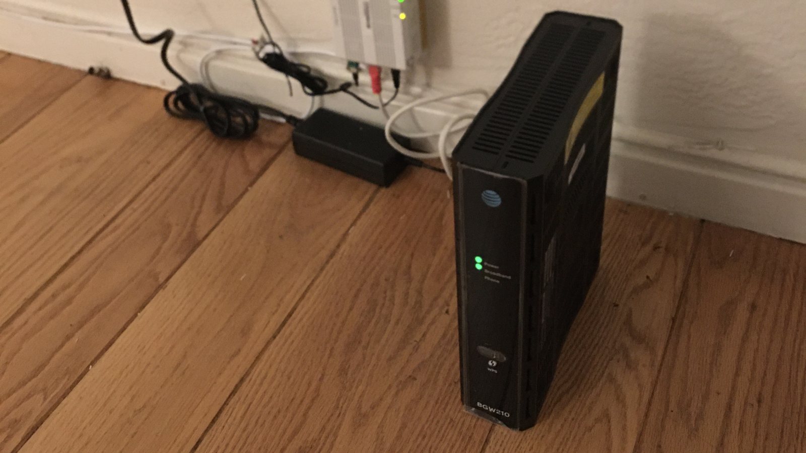 An AT&T BGW210 Wi-Fi gateway standing on a wood floor next to a wall outlet, with its WPS button on the front