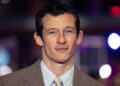 Callum Turner, in a brown suit and light blue shirt, smiling on the red carpet at the Berlin film festival