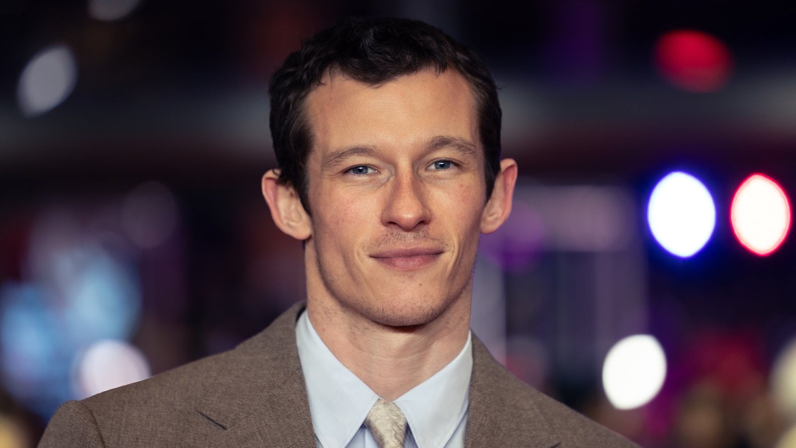 Callum Turner, in a brown suit and light blue shirt, smiling on the red carpet at the Berlin film festival