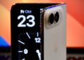 A half-open iPhone Duo standing on a desk, its inner screen showing the date Friday, October 23, in front of a laptop
