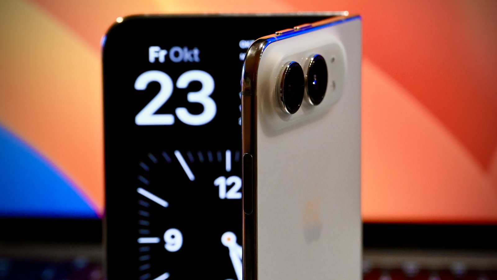 A half-open iPhone Duo standing on a desk, its inner screen showing the date Friday, October 23, in front of a laptop