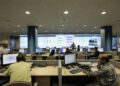 Staff at desks with computer monitors in front of a wall of large screens showing network charts in a telecom operations center