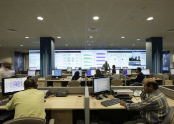 Staff at desks with computer monitors in front of a wall of large screens showing network charts in a telecom operations center