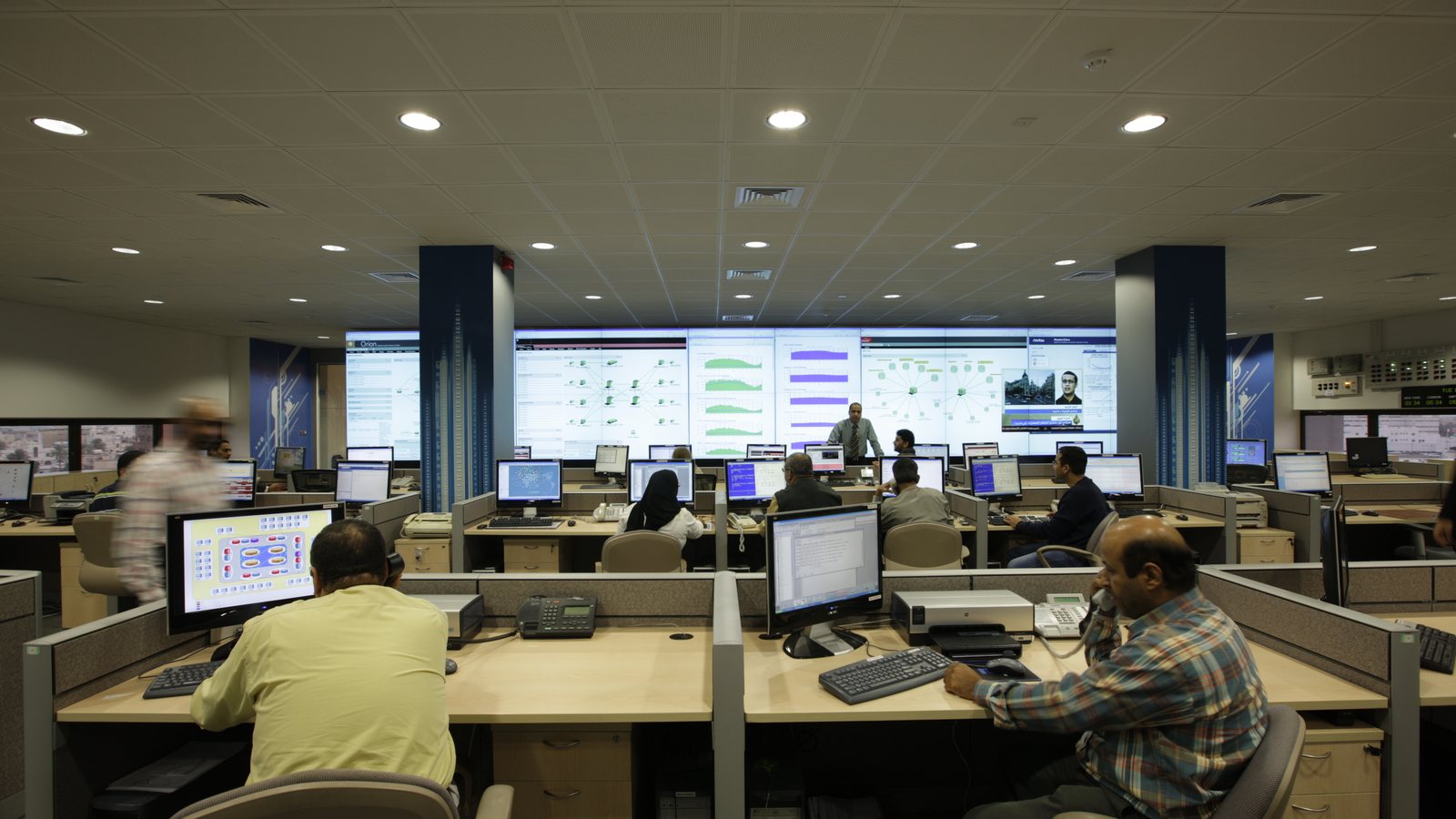Staff at desks with computer monitors in front of a wall of large screens showing network charts in a telecom operations center