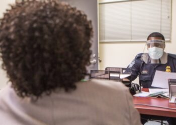 A US Customs and Border Protection officer in a face shield and mask interviews an arriving passenger at a desk at Washington Dulles airport