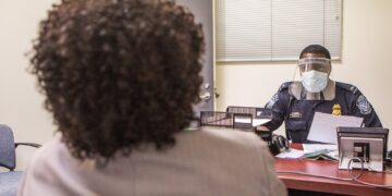 A US Customs and Border Protection officer in a face shield and mask interviews an arriving passenger at a desk at Washington Dulles airport