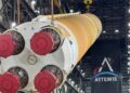 The four engine nozzles of NASA's orange Artemis III SLS core stage, covered in red caps, as cranes lift the stage inside the Vehicle Assembly Building under an Artemis banner